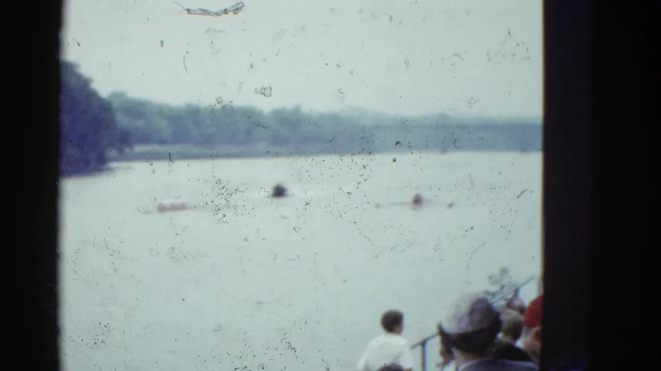 1975 Boston riverside frame showing spectators at the shore looking over the water toward distant rowers and a treelined bank.