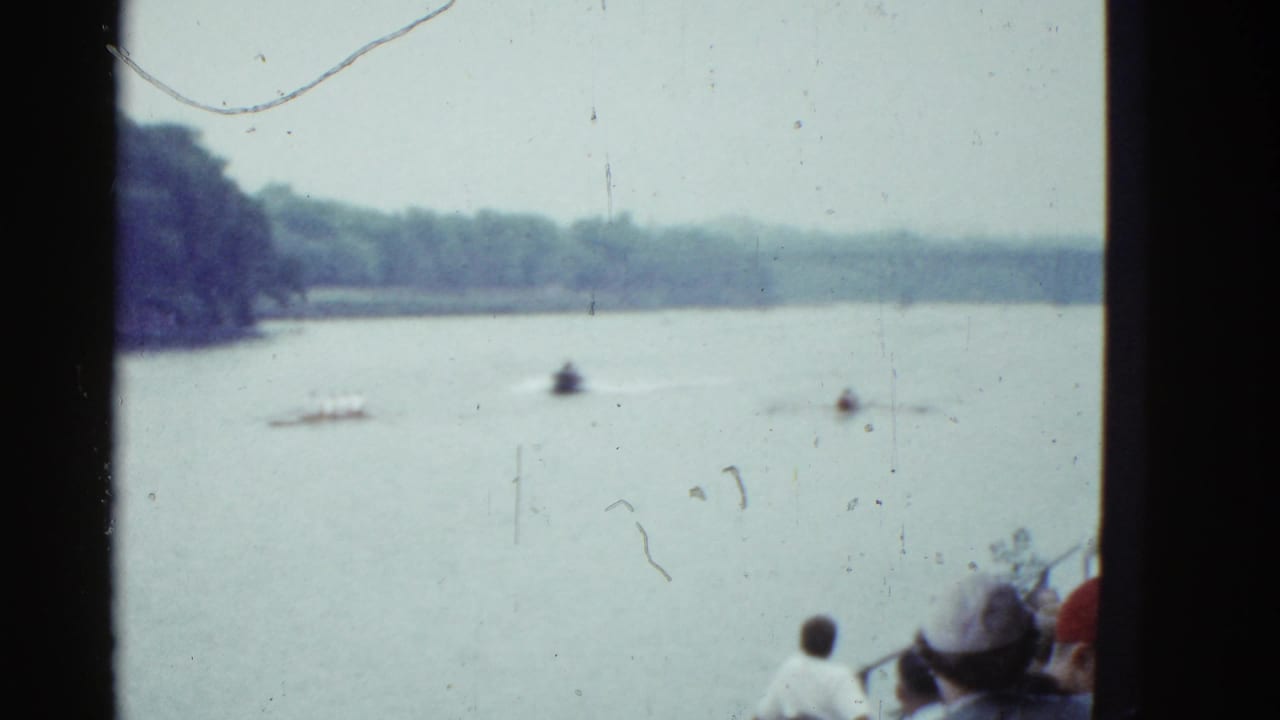 Small boats move across a broad lake with wooded banks and spectators partly visible at the lower edge.