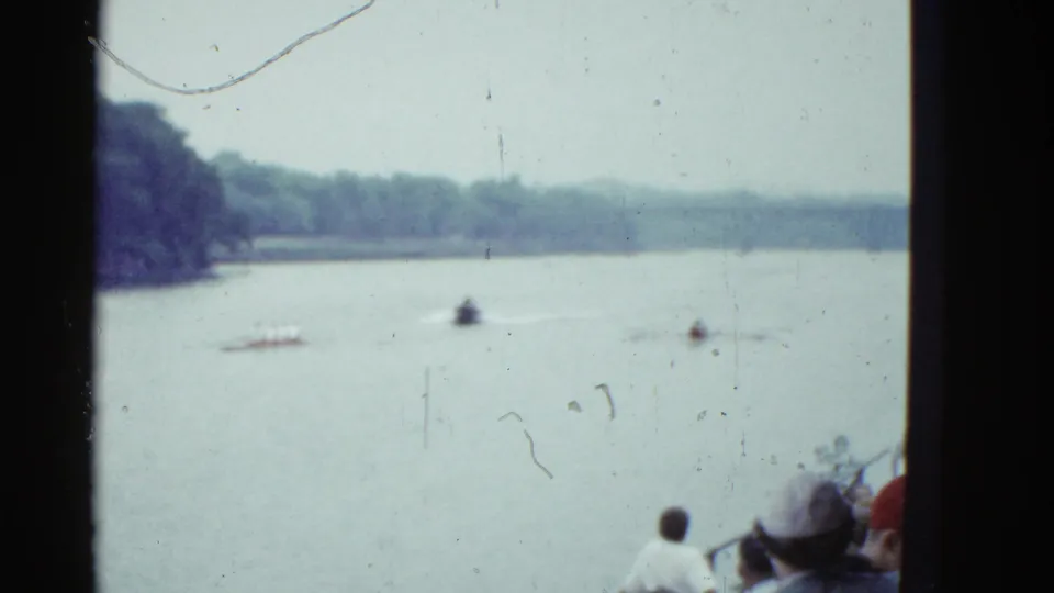 1975 8mm lakeside view near Boston, with a motorboat, rowboat, wooded shoreline, and partially visible spectators in the foreground.