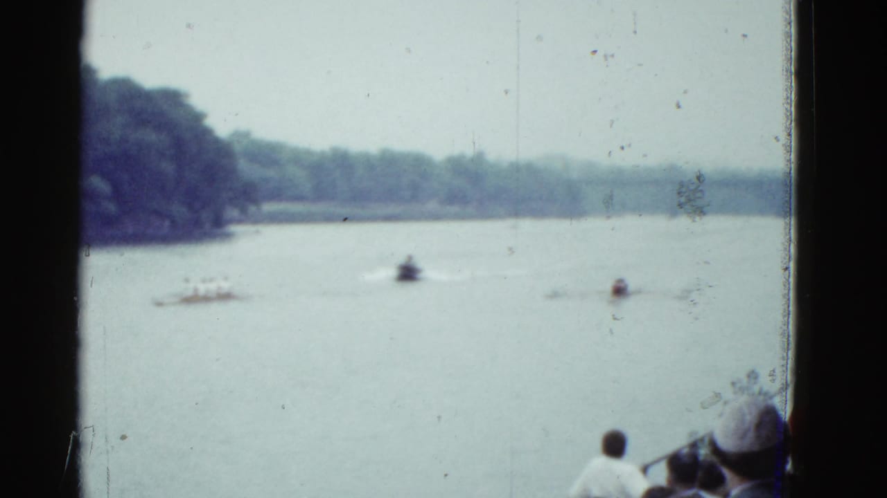 Soft 8mm frame of boats on a wooded lake near Boston in 1975, with a few foreground viewers partly visible.