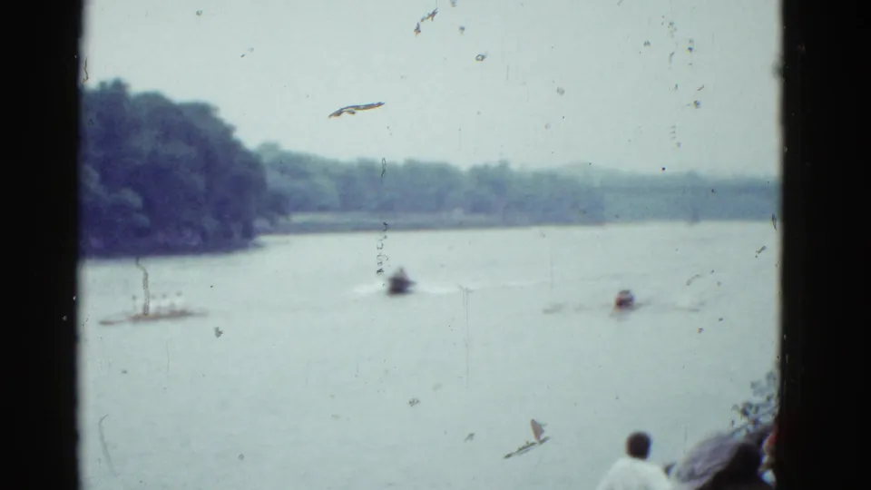 1975 home-movie frame near Boston showing small boats on a quiet lake, wooded shoreline, and a partially visible spectator below.