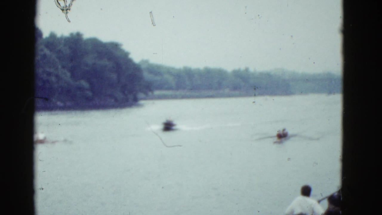 1975 Boston-area 8mm frame showing a rowing shell and motorboat on a lake, with wooded shoreline and a boat-side spectator in the foreground.
