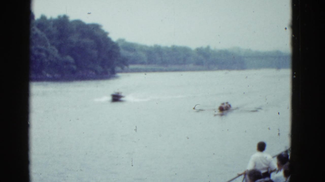 Motorboat and rowing shell move across a lake with wooded shoreline and foreground boat passengers at right.