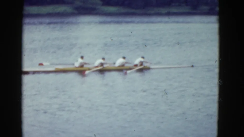1975 Boston-area 8mm frame with four rowers in white shirts moving across a lake in a long racing shell.