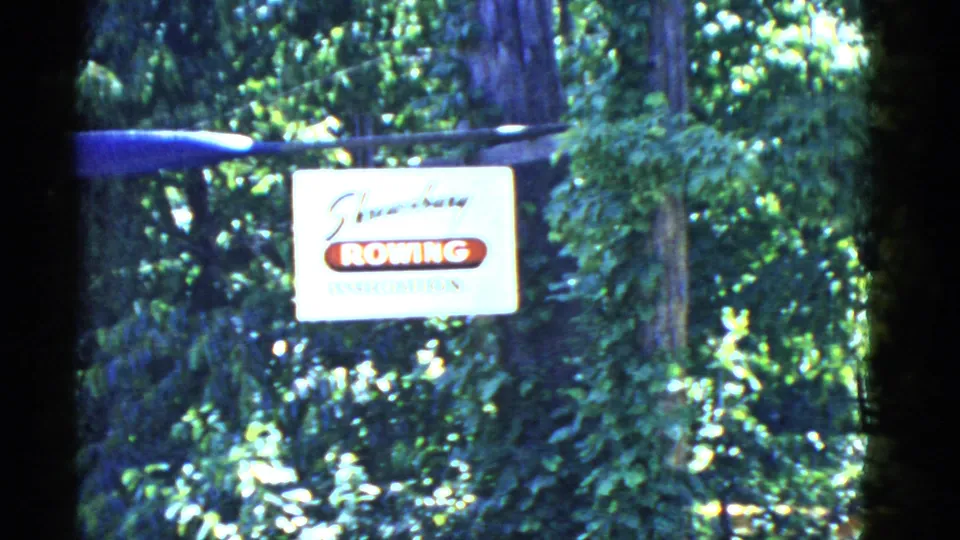 Daytime 8mm frame showing a Rowing Association sign on a fence amid dense green trees in Boston-area footage.