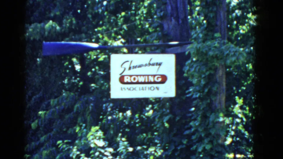 1975 8mm frame from Massachusetts showing a Shrewsbury Rowing Association sign in foliage with a blue oar mounted overhead.