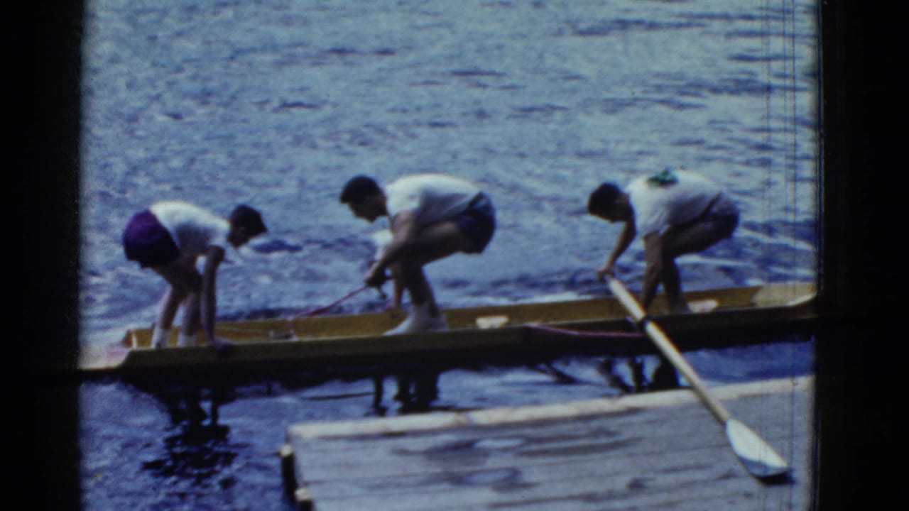 Three young men steady a narrow wooden boat beside a dock on calm water.