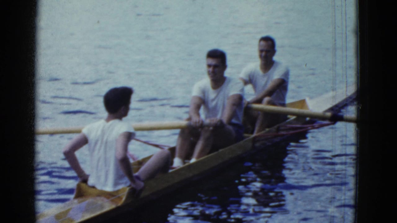 1975 Boston-area lake footage showing three young men in a narrow wooden boat, with oars extending over the water.