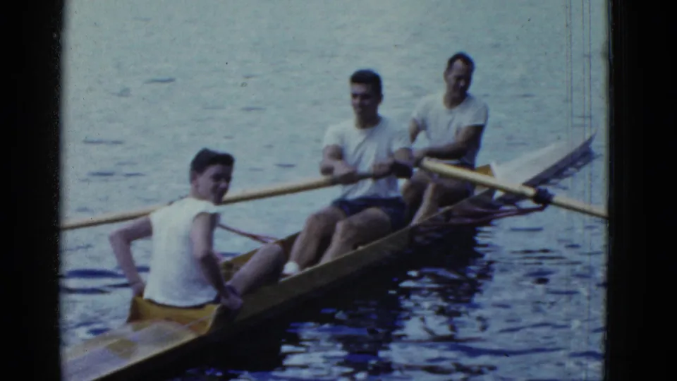 1975 Boston lake home-movie frame with one young man turning toward the camera and two others seated with oars behind him.