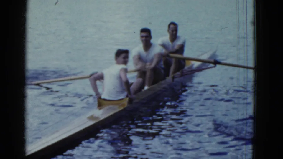 1975 Boston-area 8mm frame showing three young men rowing a narrow wooden boat across a calm lake.