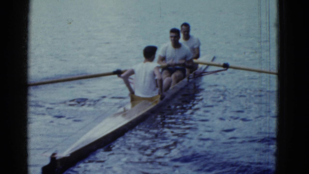 Three men row a narrow racing shell toward the camera on calm lake water.