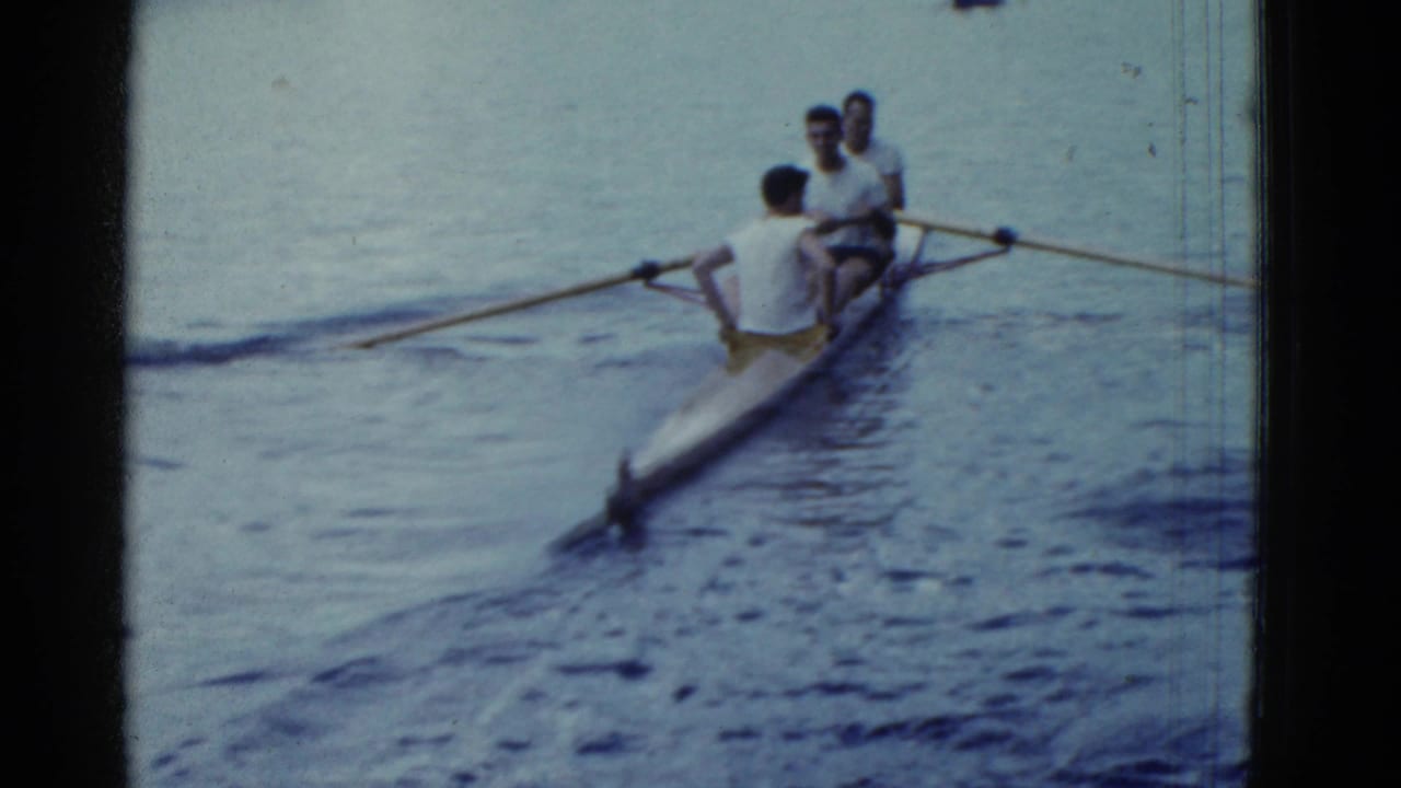 The three-man rowing shell begins to move away across placid water in soft 1975 8mm footage.