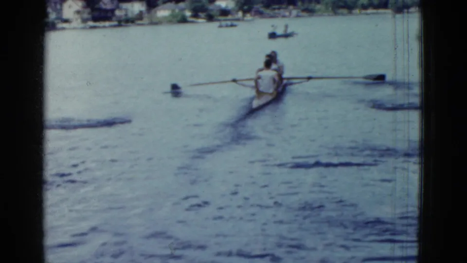The rowing shell recedes across the lake, with distant boats and a faint shoreline visible in the 1975 home movie frame.