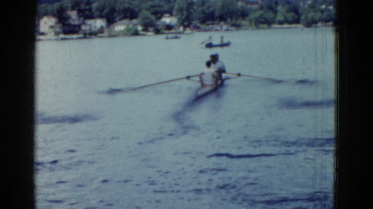 1975 Boston-area 8mm frame showing two men in a rowboat on calm water, with a wooded far shore and small boats in the distance.