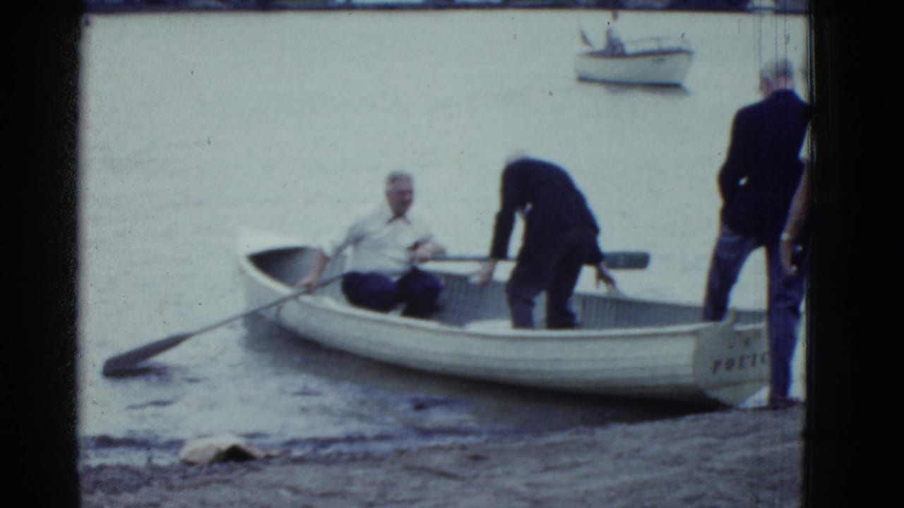An older man sits in a white rowboat while two men stand near the lakeshore.