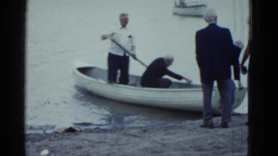 1975 Boston-area 8mm frame showing an older man standing with an oar in a white rowboat near shore as others remain nearby.