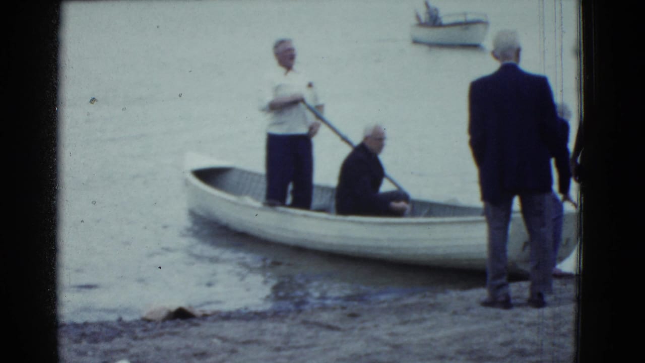 1975 Boston-area 8mm frame showing adult men preparing a small white rowboat at a calm shoreline.