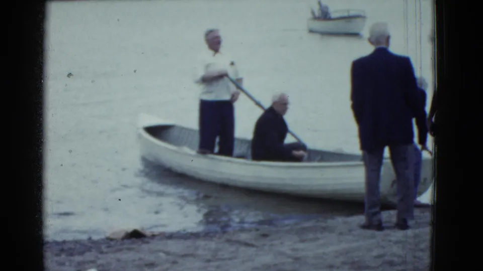 1975 Boston-area 8mm frame showing adult men preparing a small white rowboat at a calm shoreline.
