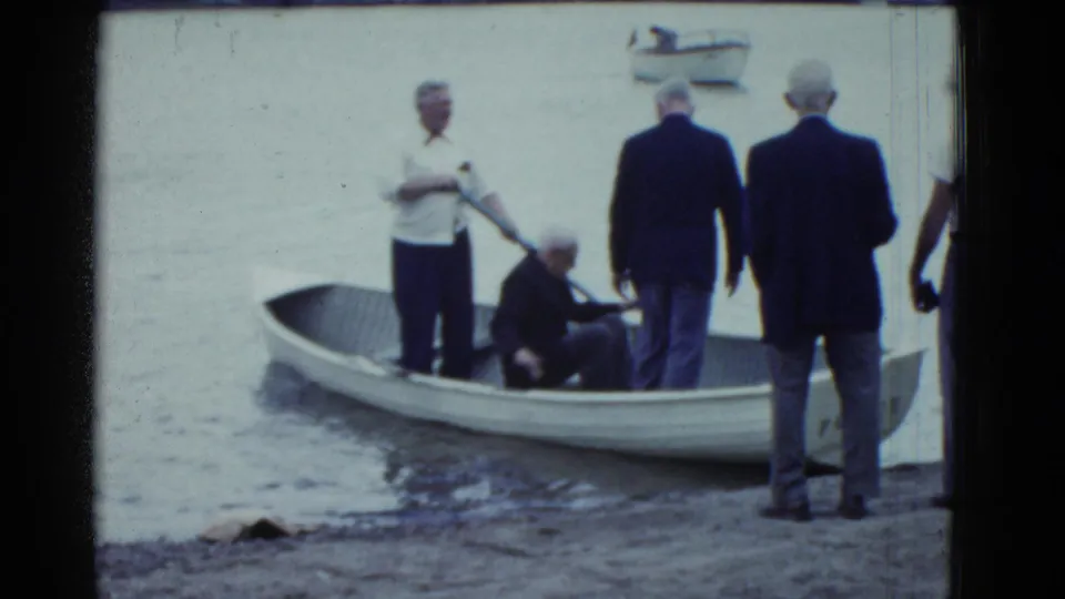 Boston-area 1975 8mm frame with men in suits near a white rowboat as boarding continues at the shoreline.
