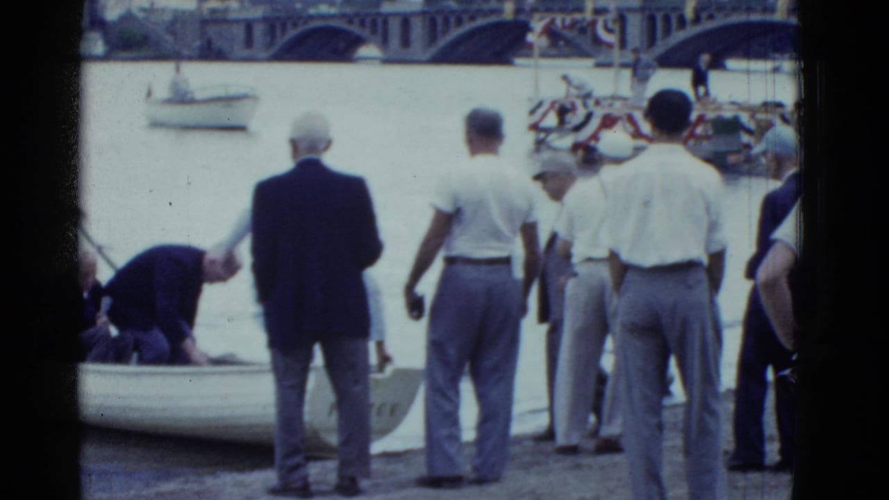 Men stand near a rowboat as a man bends over at the river edge.