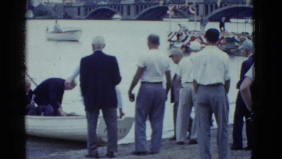Older men in light shirts and slacks stand near a small rowboat on the Boston riverfront, with a bridge and boats behind them.