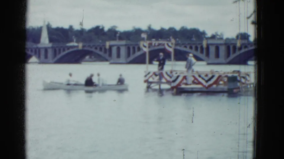 Wide 1975 Boston river view with a small rowboat, decorated floating platform, and large arched bridge in the background.