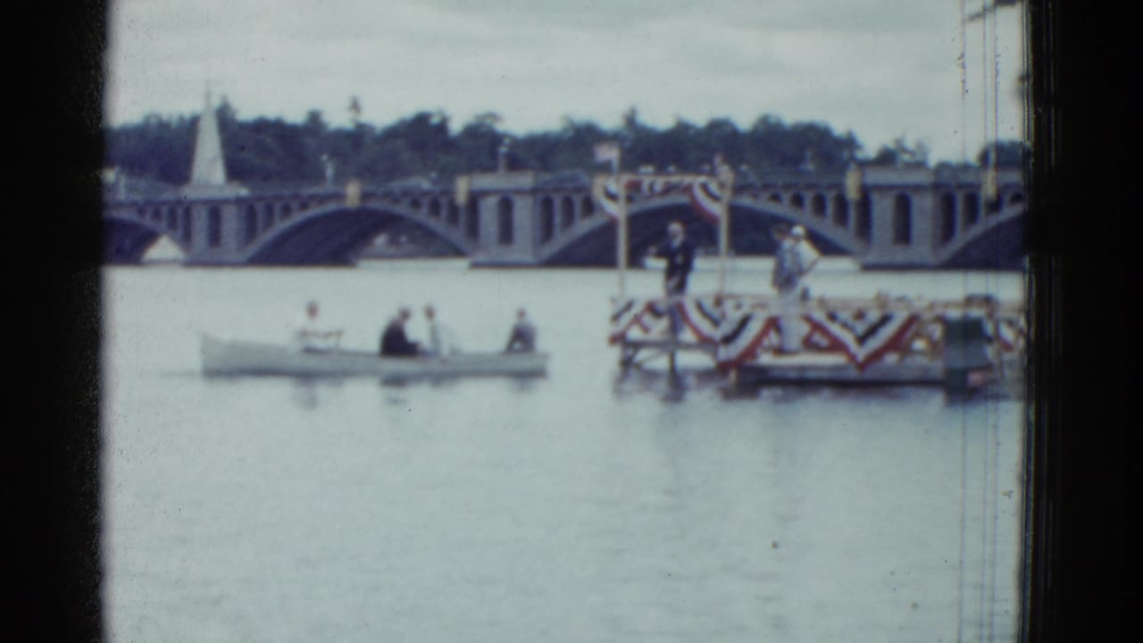 1975 Boston home-movie frame showing adults in a small boat near a decorated floating platform, with an arched bridge in the background.