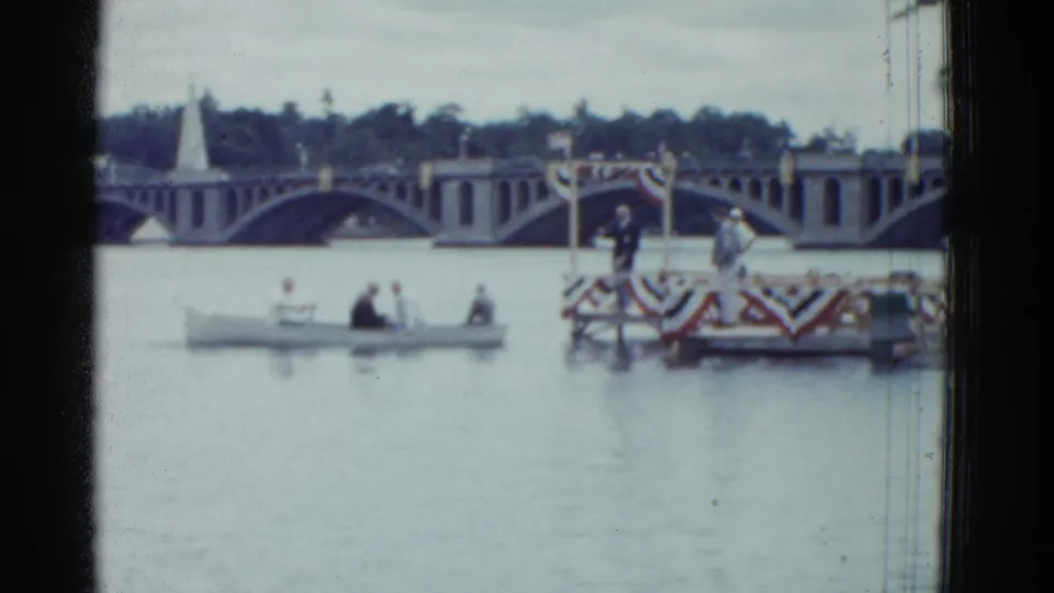 1975 Boston home-movie frame showing adults in a small boat near a decorated floating platform, with an arched bridge in the background.