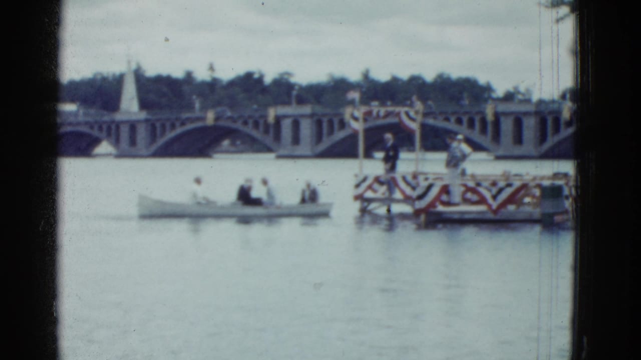 Boat with several adults moves beside a decorated platform on a river.