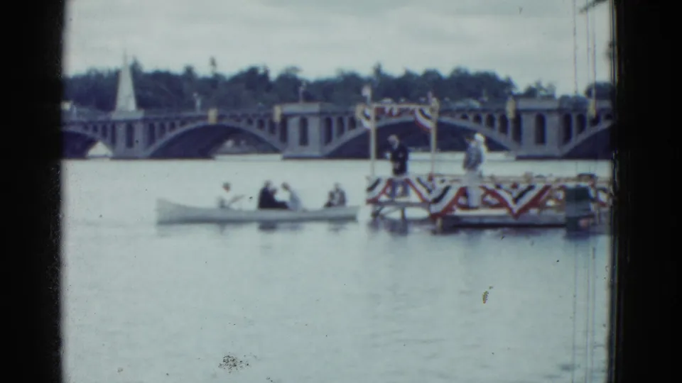 1975 Boston river scene with adults in a small boat passing in front of a patriotic-looking platform and arched bridge.
