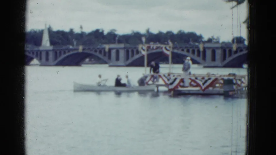 1975 Boston home-movie frame showing a bunting-draped boat and rowboat on a river before an arched bridge.