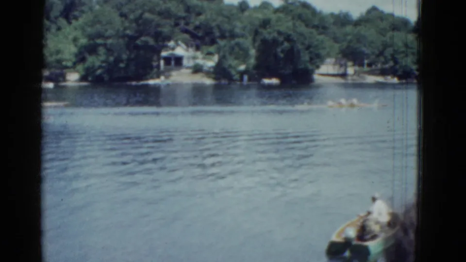 1975 8mm river scene in Boston with rowing shells in the distance and a small foreground boat carrying a rower at the edge of frame.