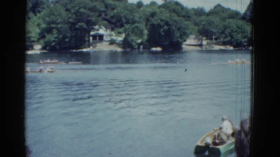 Boston-area 1975 8mm frame with several rowing boats on calm water, wooded shore, and a foreground man in a small green boat.