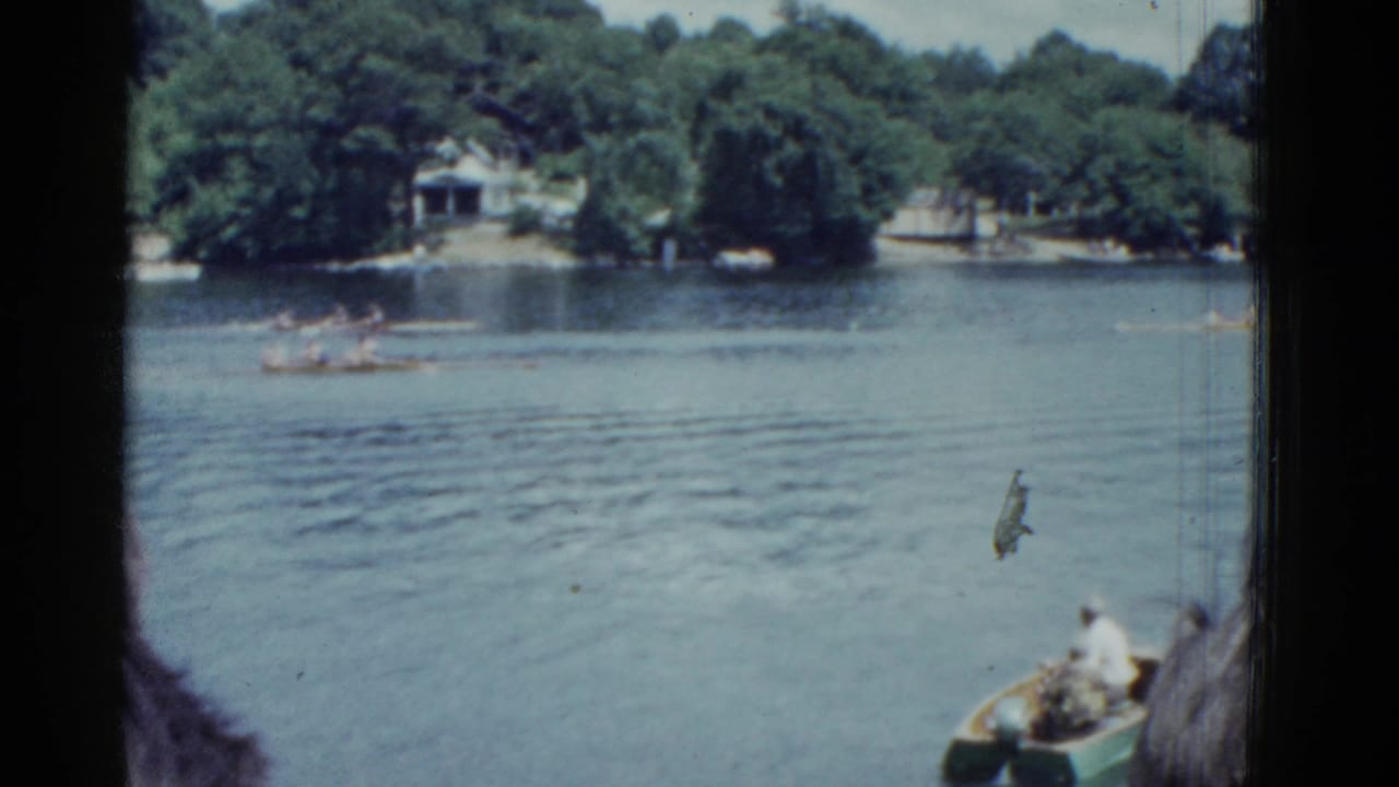Two rowing shells move across a lake with trees and houses along the opposite shore.