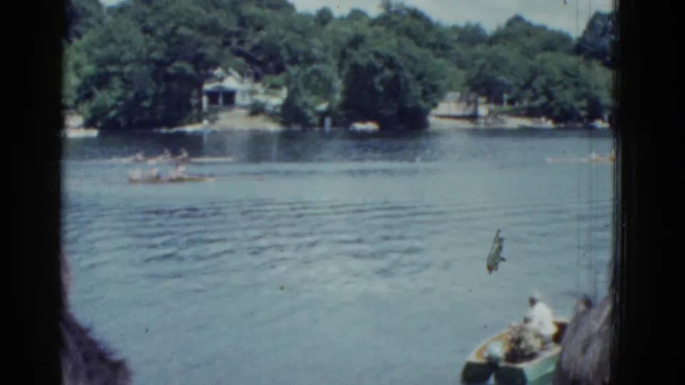 1975 Boston 8mm footage shows rowing shells on a lake, viewed from near another boat with a wooded shoreline behind them.