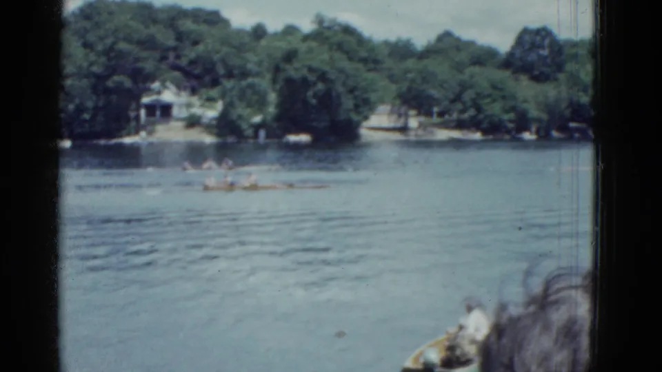 A mid-1970s home-movie frame shows rowing shells moving across a Boston lake with houses and trees in the background.