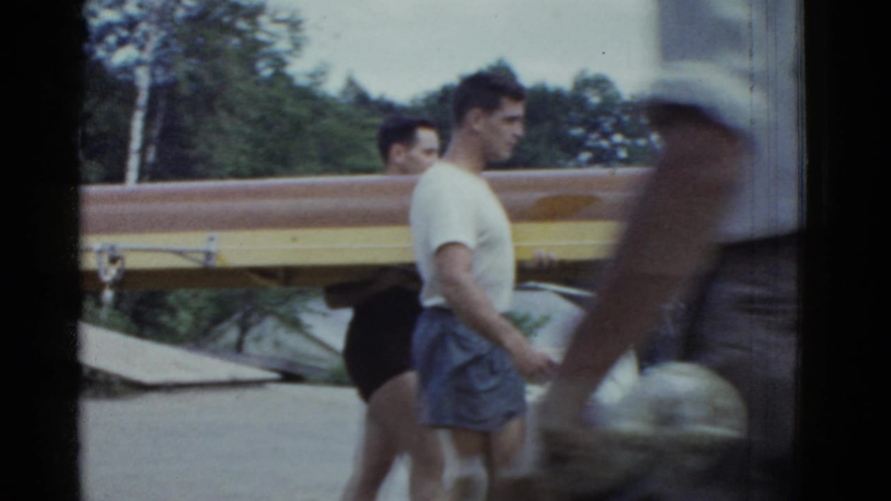 Adult rowers carry a long wooden shell beside a lake while a blurred person passes in front.