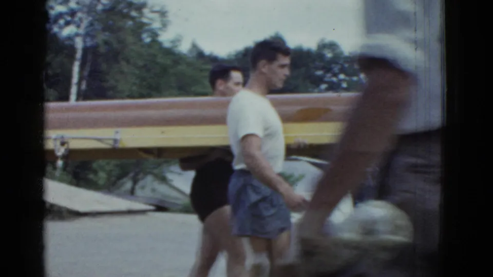 In 1975 Boston-area 8mm footage, adult male rowers carry a long wooden racing shell near lakeside water and trees.