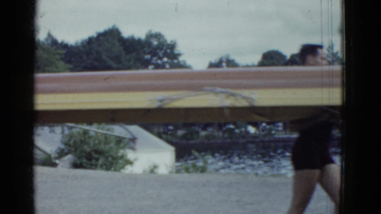 The wooden shell fills the frame as rowers move past the camera near a lakeside setting in Boston-area 1975 footage.