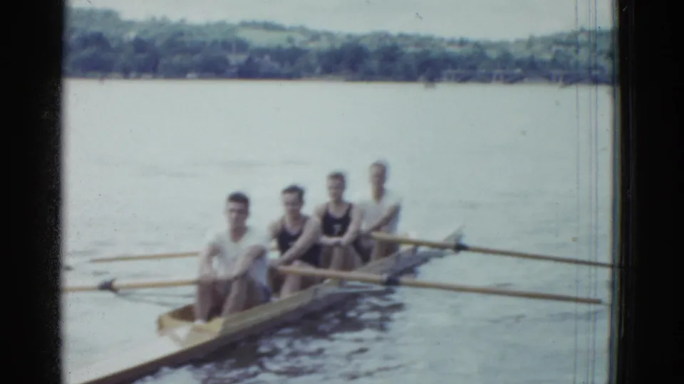 Four adult male rowers sit in a narrow racing shell on calm water, oars out, with a wooded shoreline behind them.