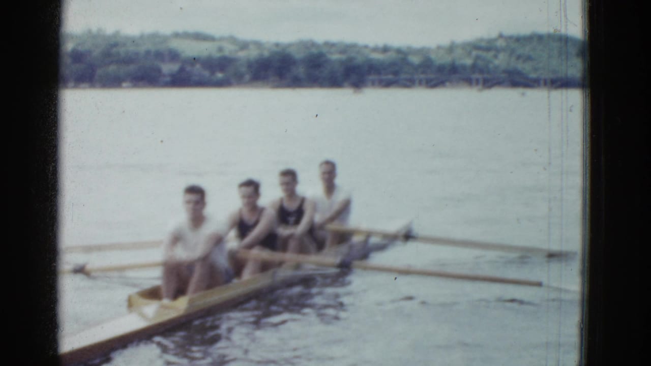 Four adult men in a narrow rowing boat appear on a calm lake in Boston-area 1975 8mm home-movie footage.