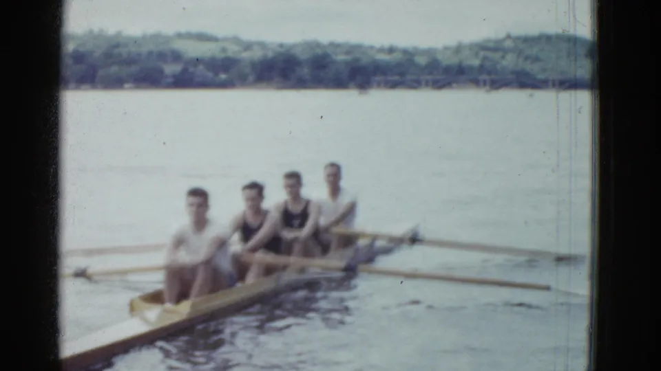 Four adult men in a narrow rowing boat appear on a calm lake in Boston-area 1975 8mm home-movie footage.