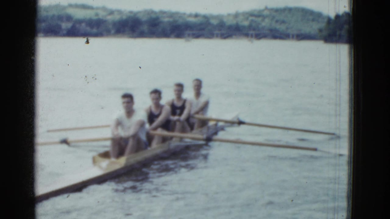 Four men face the camera from a rowing boat with oars spread across the water.