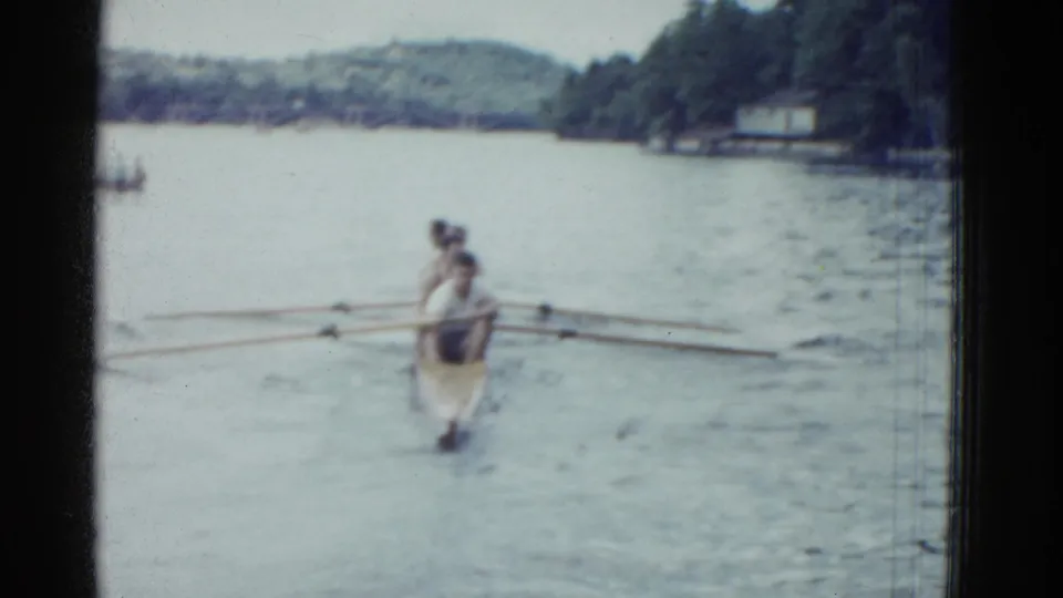 The narrow rowboat moves farther from the camera on a lake, with wooded shore and distant structures visible in 1975 home-movie footage.