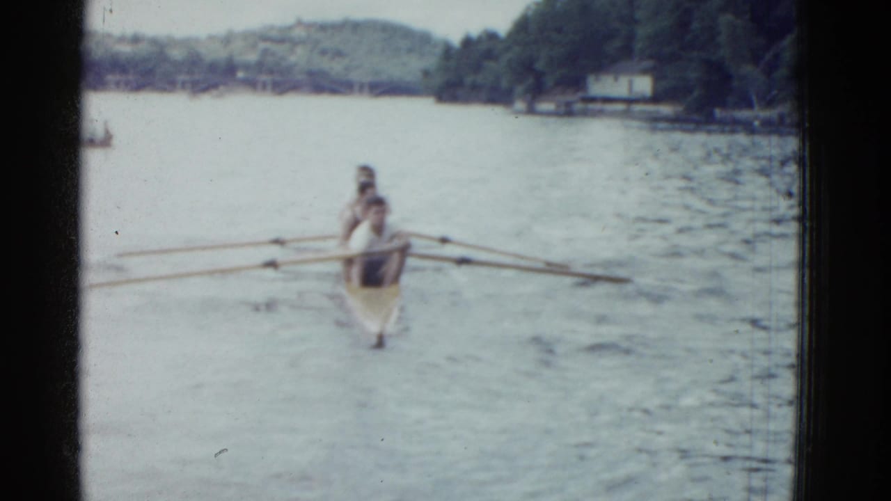 Two rowers approach the camera in a narrow shell on a lake, with oars spread across the water.