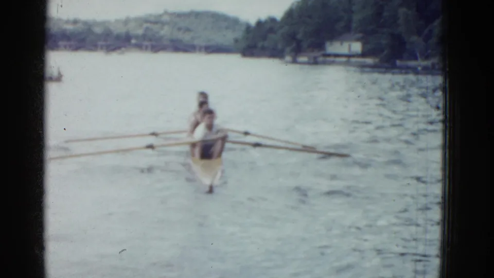 1975 Boston-area 8mm frame showing rowers in a narrow shell approaching across a calm lake with wooded shoreline behind them.