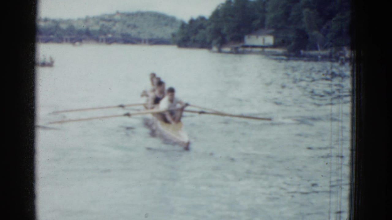 Rowing shell draws closer on the lake, with several men seated in line and oars stretched outward.