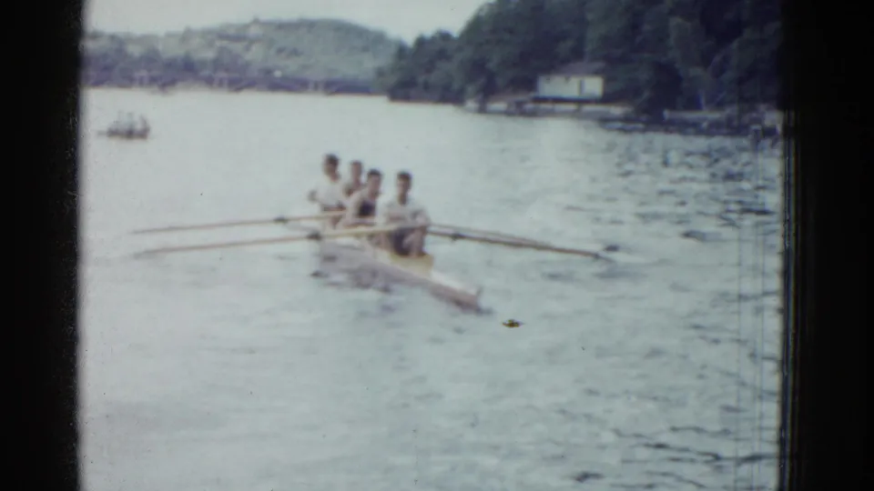 Four adult men row in unison on a lake in 1975 Boston-area home-movie footage, with a wooded shoreline and distant hills visible.
