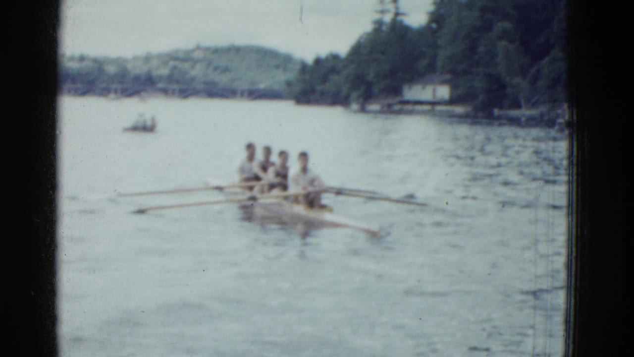 1975 Boston-area 8mm frame showing four men rowing a shell on a calm lake with trees, distant hills, and a small shoreline building.