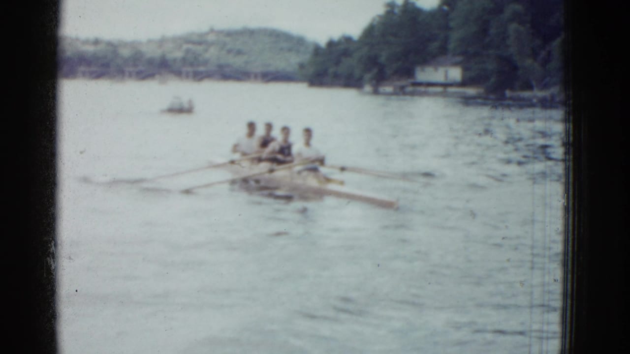 The crew rows in unison across a Boston-area lake in soft 8mm footage, with a wooded bank and low hills in the background.